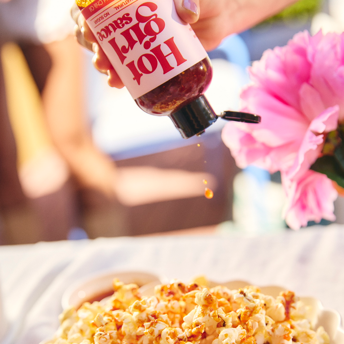 Woman squeezing chili oil from a bottle of OG Hot Girl Squeezable Chili Oil into a bowl of popcorn at a summer backyard picnic - Hotgirlsauce chili oil - chili sauce - hot sauce
