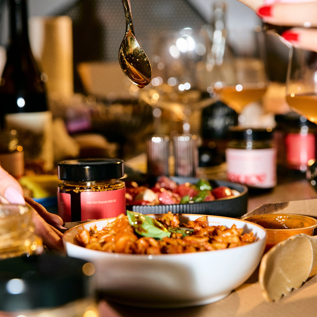 Spoon dripping chili oil into a bowl of pasta on a table surrounded with Hotgirlsauce chili oil and Italian dishes -  Hotgirlsauce chili oil - chili sauce - hot sauce