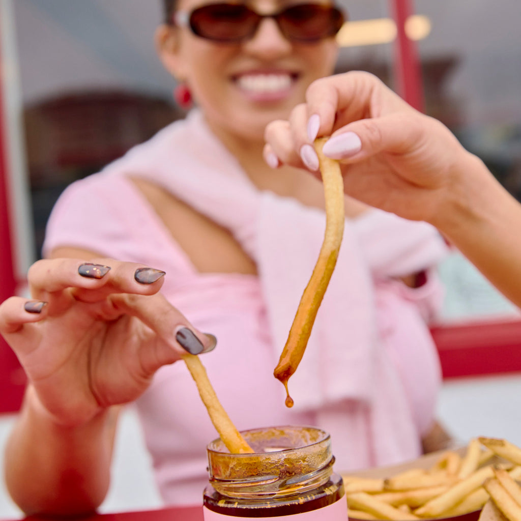 Woman smiling and dipping a fry into a jar of OG Hot Girl Chili Oil hot sauce in casual dining setting -  Hotgirlsauce chili oil - chili sauce - hot sauce