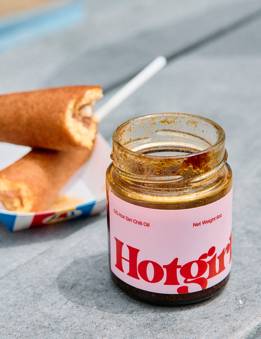 Jar of OG Hot Girl Chili Oil next to a couple of corn dogs on a gray picnic table  -  Hotgirlsauce chili oil - chili sauce - hot sauce