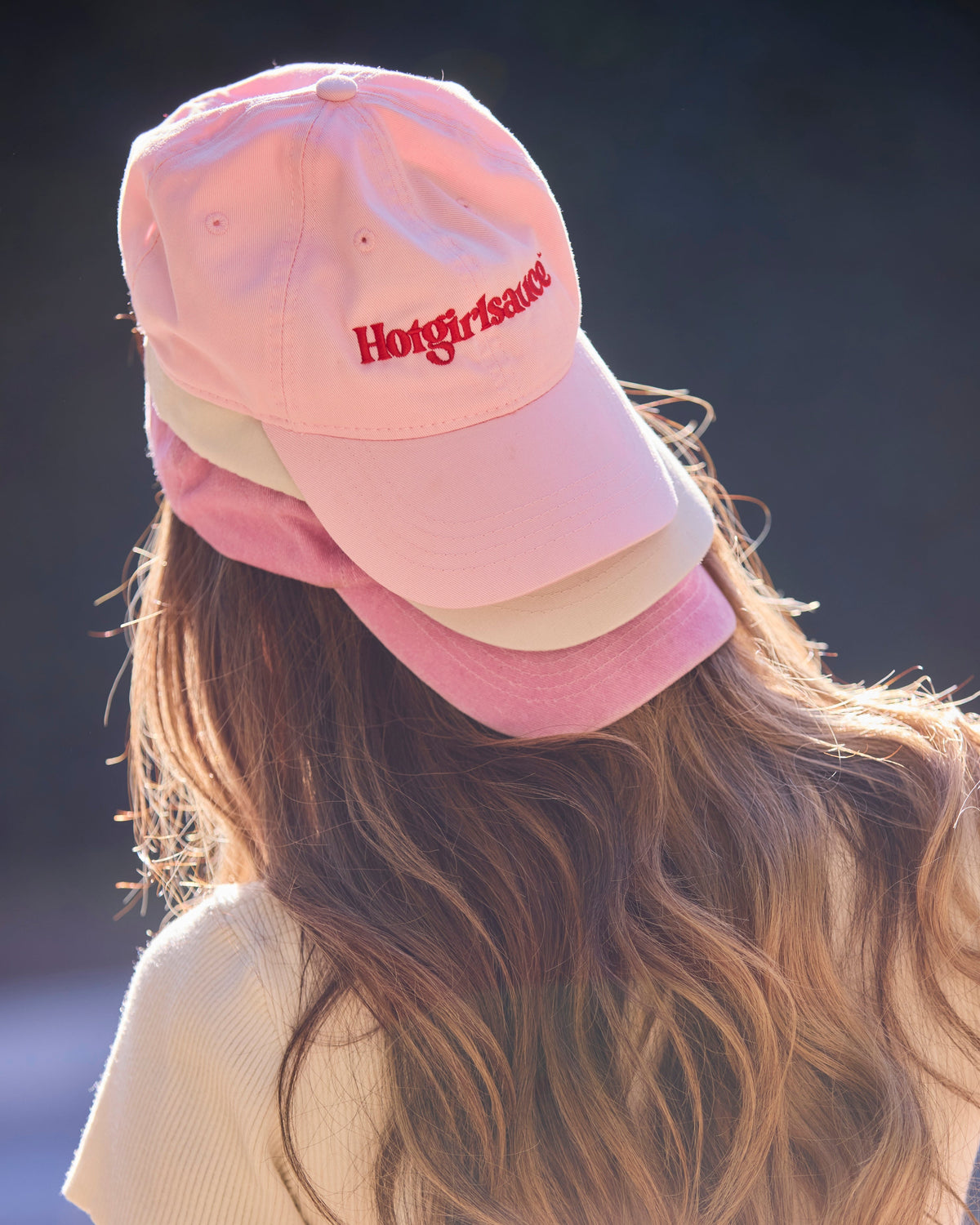 Back view of blush pink Hotgirlsauce dad hat with embroidered logo, worn outdoors, stacked on other hats