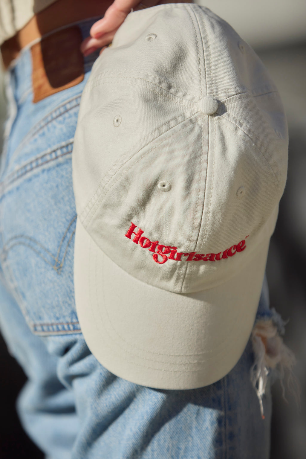 Close-up of Hotgirlsauce embroidered logo on cream dad hat in hands by hips