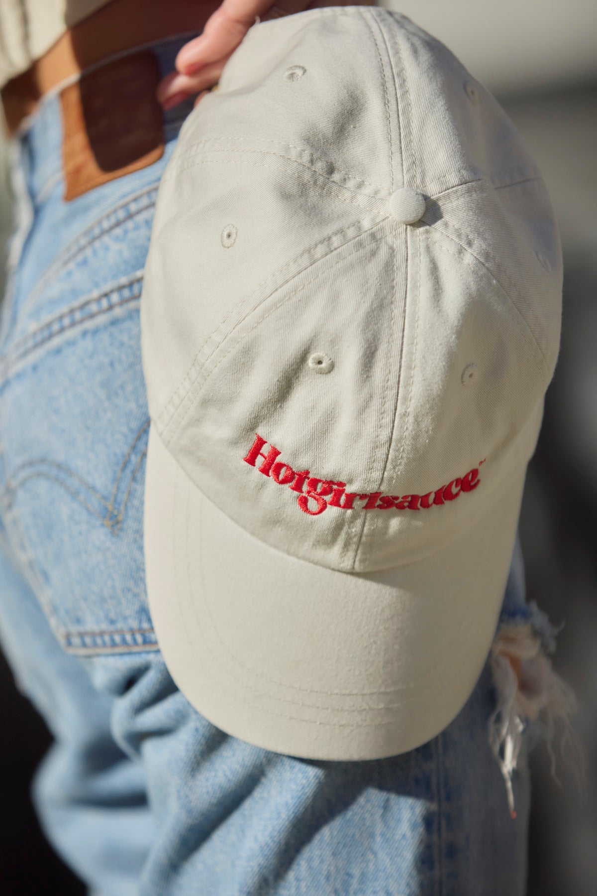 Close-up of Hotgirlsauce embroidered logo on cream dad hat in hands by hips