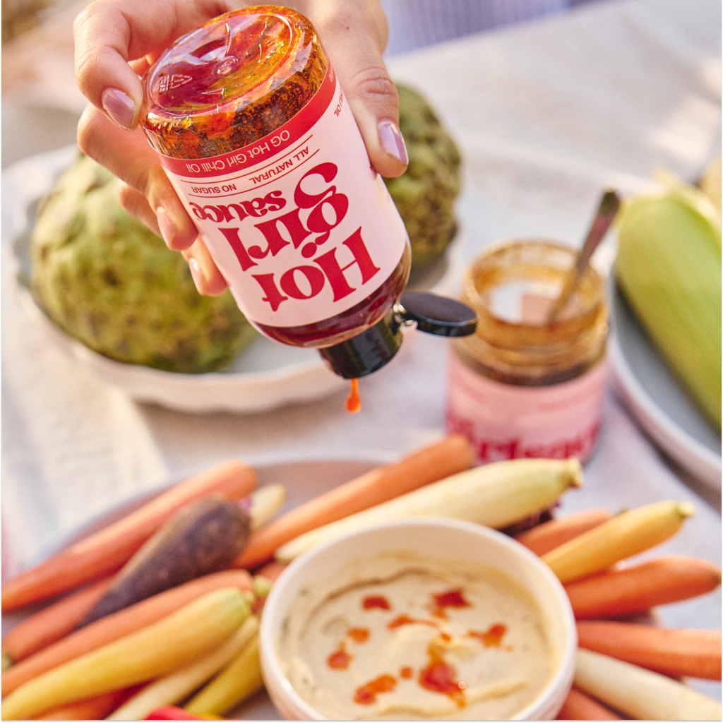 Woman squeezing OG Hot Girl Chili Oil into bowl of hummus during summer backyard picnic -  Hotgirlsauce chili oil - chili sauce - hot sauce