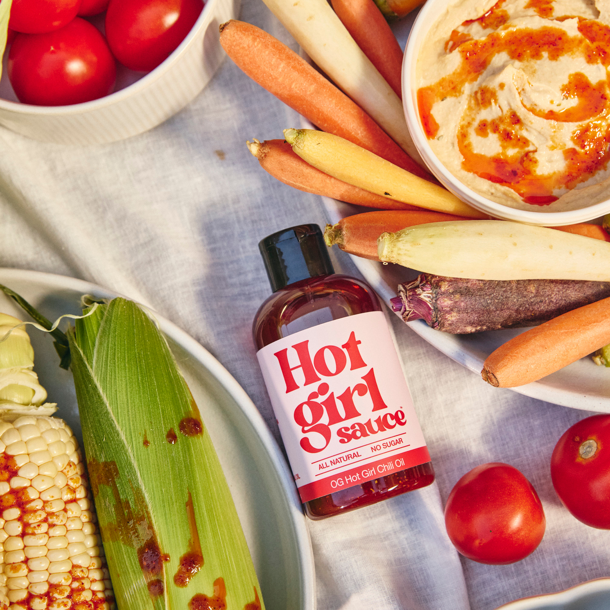 Bottle of OG Hot Girl Squeezable Chili Oil lying next to hummus, carrots, corn on the cob, and tomatoes at a summer picnic - Hotgirlsauce chili oil - chili sauce - hot sauce